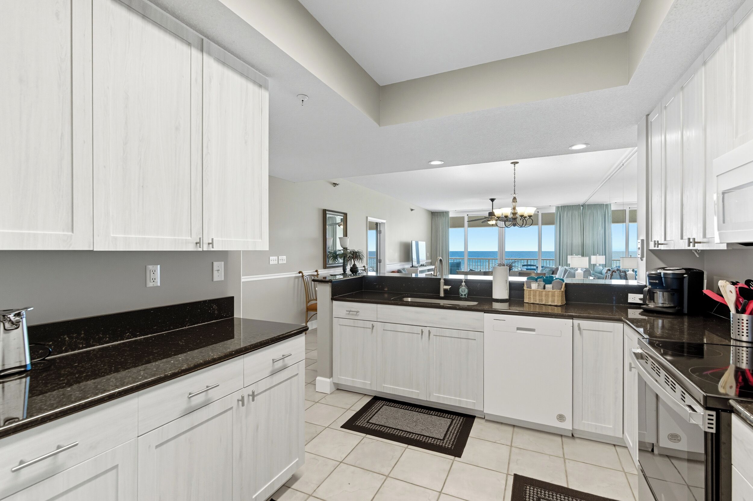 A modern kitchen with white cabinets, black countertops, and a large island. There is a view of the ocean through the windows.