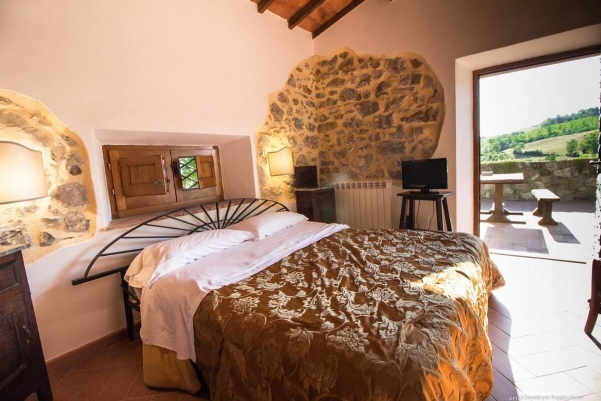 Une chambre avec un mur en pierre, un lit recouvert d’une couvre-lit à motifs, une télévision et une vue sur un paysage verdoyant.