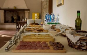 A buffet table with various food items, including meats and cheeses, in a room with a fireplace and a yellow lamp in the background.