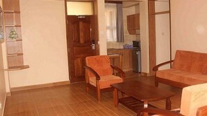 Una sala de estar con un sofá, dos sillones y una mesa de centro. Se ve una puerta de madera y una cocina al fondo.