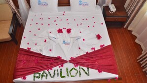 Una cama bien hecha con una colcha roja y blanca, almohadas con el logotipo de 'Pavilion Holiday Resort' y un arreglo decorativo de flores y toallas.