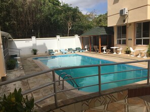 Una piscina con sillas de descanso y mesas, rodeada de un muro blanco y árboles.
