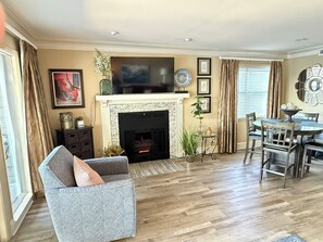 A living room with a fireplace, a television, a dining area with a table and chairs, and a large window with curtains.
