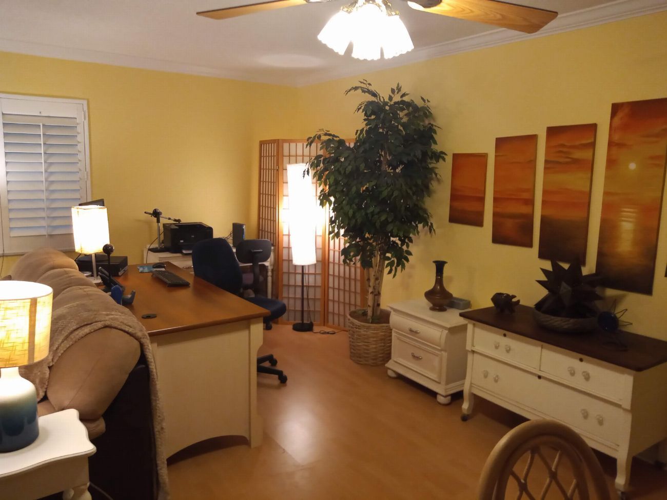 A well-lit living room with a desk, a chair, a sofa, a plant, and a wheelchair.