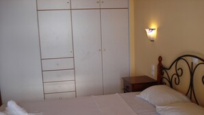A neatly made bed with white linens and towels, a wooden nightstand, a wall-mounted light, and a wardrobe with multiple drawers. - Βίλα Μιχάλης  πανόραμα (Kombi)