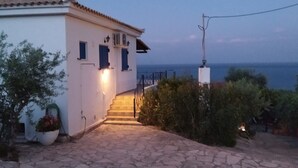 A white building with a tiled roof, a stone staircase leading to a door, and a view of the sea in the background. - Βίλα Μιχάλης  πανόραμα (Kombi)