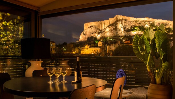 A table with wine glasses and a bottle, a potted plant, and a view of a historic building at night. - Fos Residential Apartments (Athens)