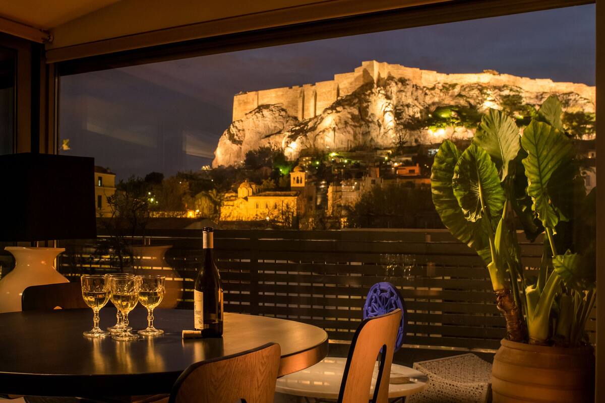 A table with wine glasses and a bottle, a potted plant, and a view of a historic building at night.