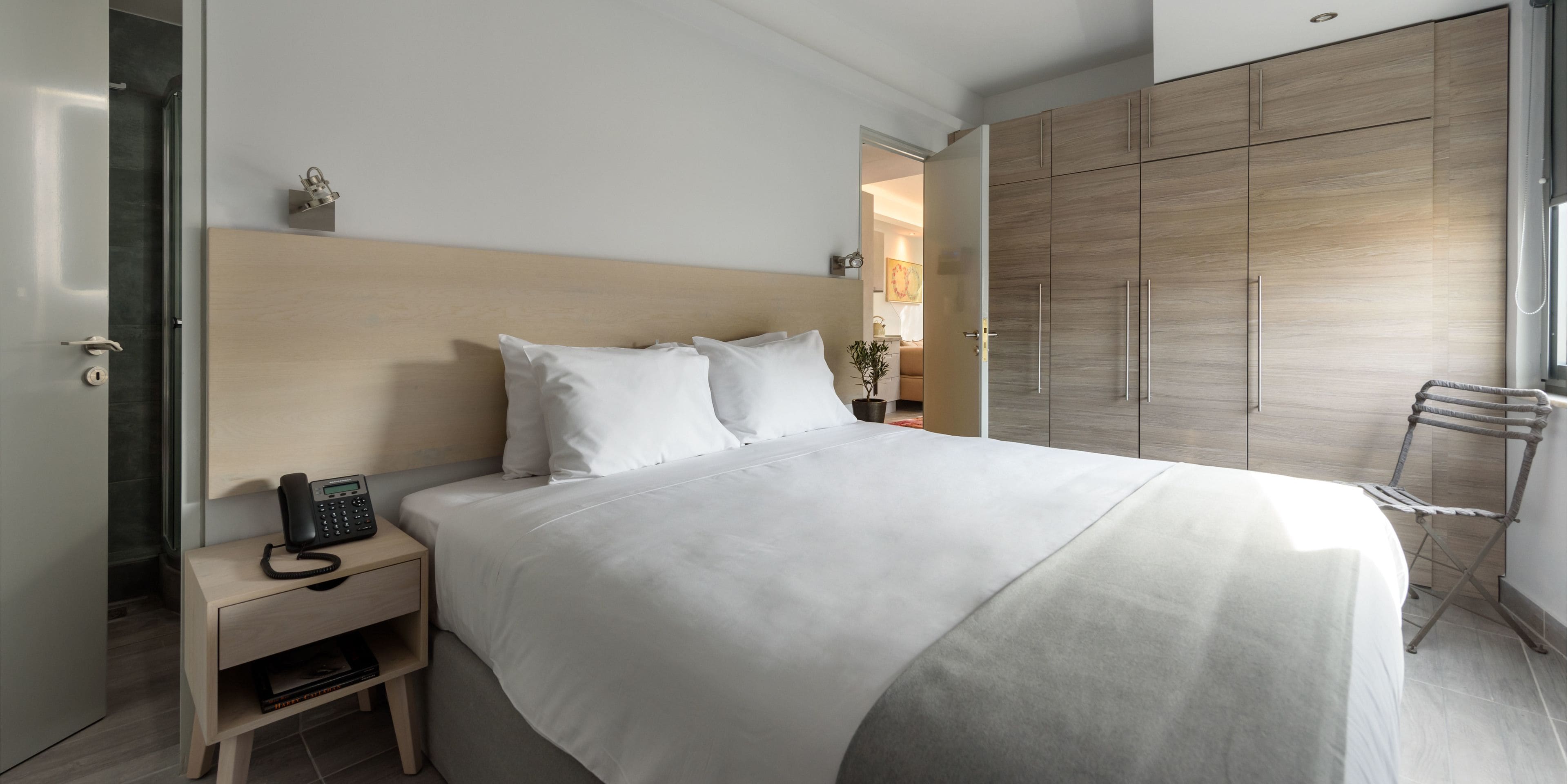 A modern hotel room with a large bed, a bedside telephone, a wooden wardrobe, and a view of the adjacent room.