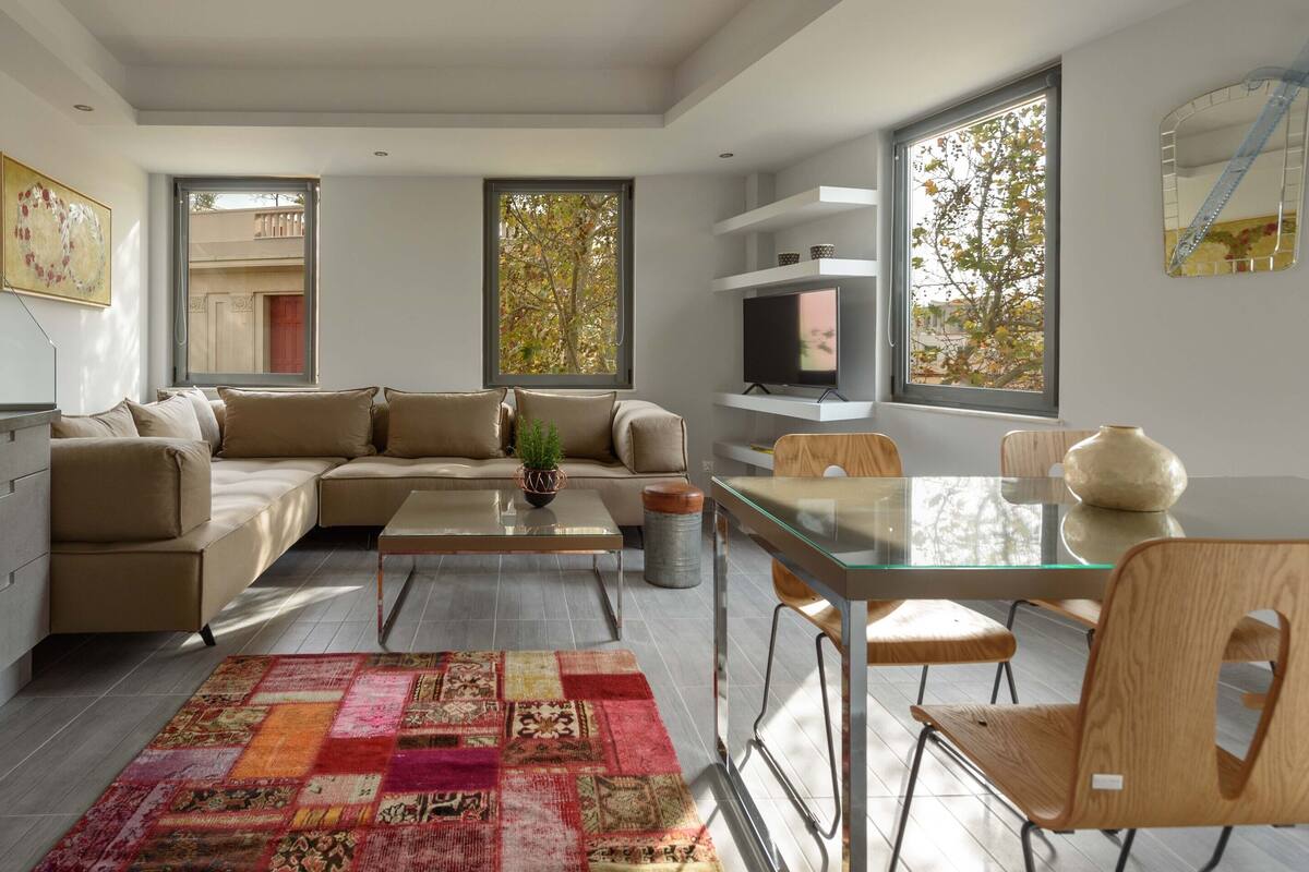 A modern kitchen with a built-in oven, a dining area with a glass table and wooden chairs, a beige sofa, and a colorful rug.