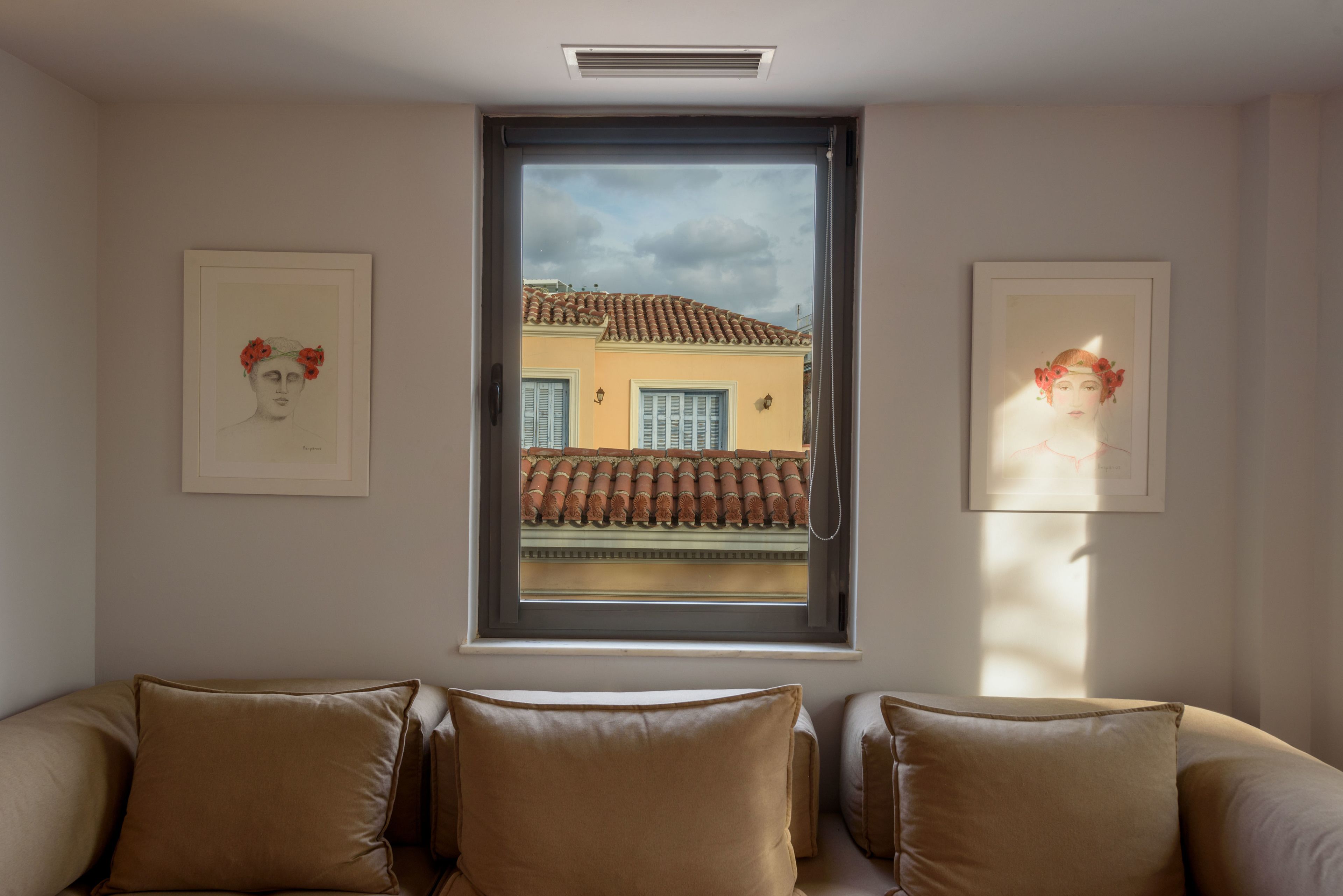 A room with a window overlooking a building with a tiled roof. There are two framed artworks on the wall, a brown leather sofa, and a small table.
