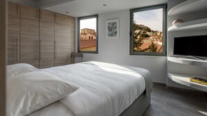 A modern bedroom with a bed, a TV, and a view of a cityscape through a window. - Fos Residential Apartments (Athens)