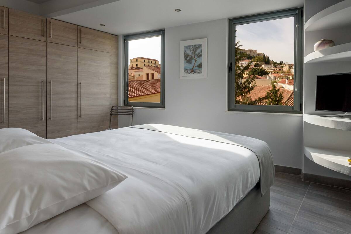 A modern bedroom with a bed, a TV, and a view of a cityscape through a window.