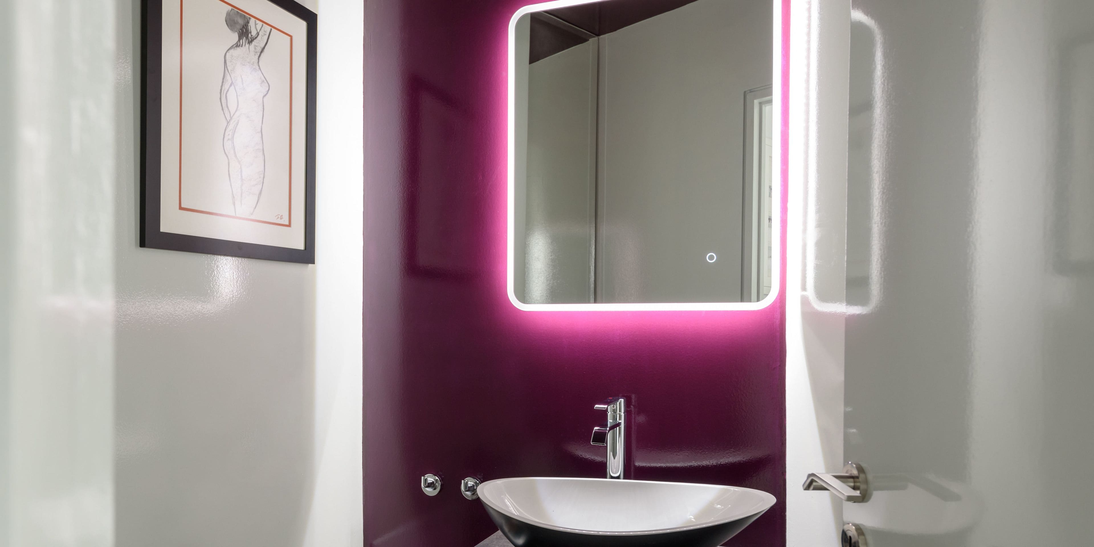 A bathroom with a white sink, a mirror with a neon light, and a framed artwork on the wall.