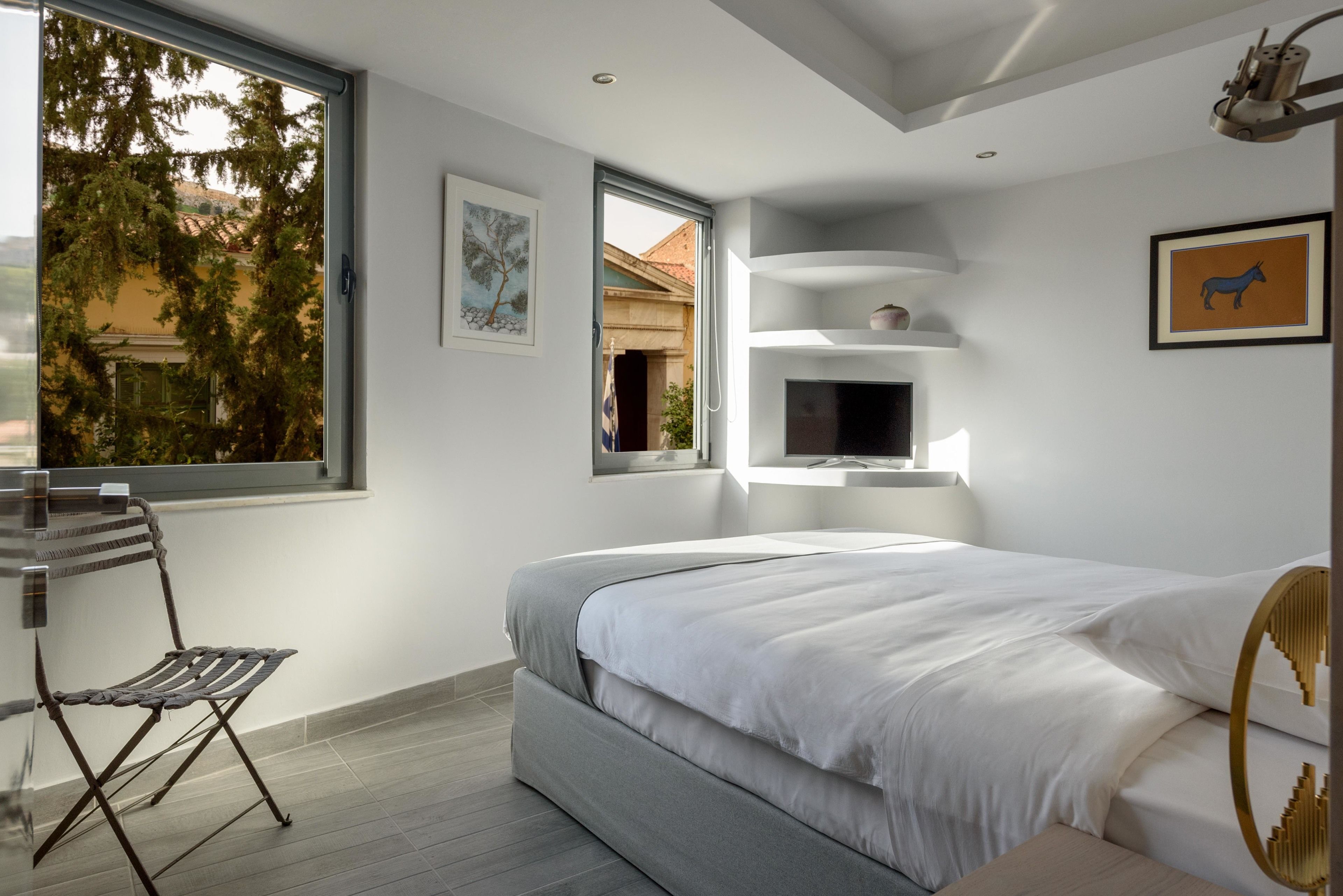 Een moderne slaapkamer met een groot bed, een stoel, een televisie en een raam met uitzicht op bomen en gebouwen.