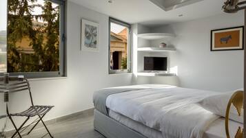 Een moderne slaapkamer met een groot bed, een stoel, een televisie en een raam met uitzicht op bomen en gebouwen.