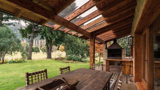 Una terraza de madera con una mesa, sillas y un banco de madera, junto a una chimenea de piedra.
