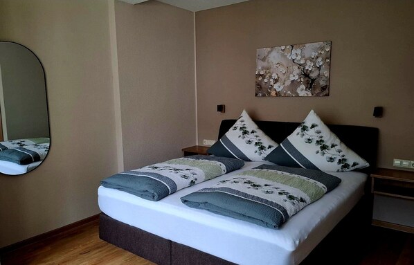 A neatly made bed with floral pillows, a bedside table, a wall-mounted mirror, and a painting above the bed. - Zum Weingutsmuseum (Cochem)