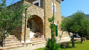 A stone building with a small balcony, a staircase leading to the entrance, and a well-maintained garden with trees and shrubs.