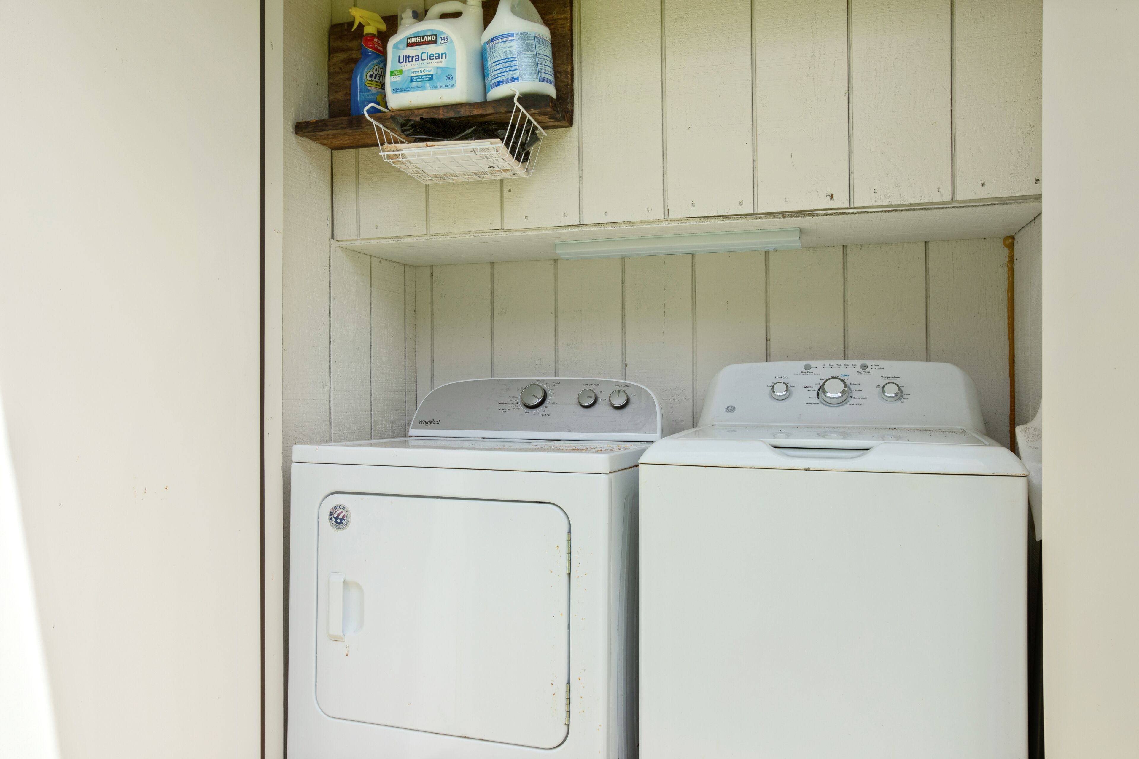 A laundry area with two white washing machines and a shelf above containing cleaning supplies.