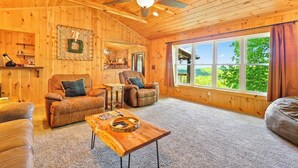 A cozy wooden cabin interior with leather sofas, a wooden coffee table, and a large window offering a view of greenery. - 'View & Far Between' Cozy Mountain Home w/ hot tub (Ashe County)