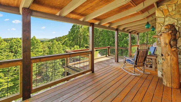 A wooden deck with a view of a forest, a stone wall, and a rustic wooden chair. - 'View & Far Between' Cozy Mountain Home w/ hot tub (Ashe County)