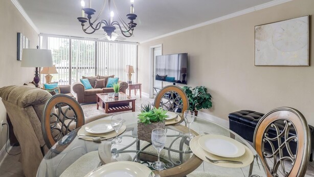 A dining area with a glass table, chairs, and a chandelier. There is a sofa and a television in the background. - Beach Days = Best Days!   Fishing, Sunning, Beaching, Boating, Swimming=FUN!! (Biloxi)