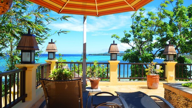 A balcony with wicker chairs, a table, potted plants, and a large striped umbrella.