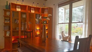 A dining area with a wooden table and chairs, a bookshelf with various items, and a view of trees through a large window.