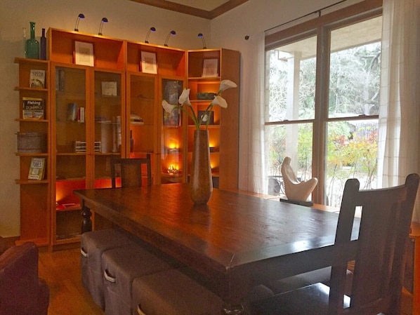 A dining area with a wooden table and chairs, a bookshelf with various items, and a view of trees through a large window.