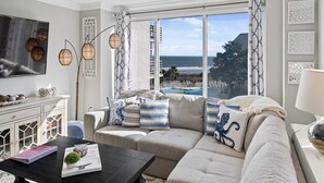 Ein modernes Wohnzimmer mit großem Fenster mit Blick auf einen Strand, eine Couch mit Zierkissen, ein Couchtisch mit Büchern und einer Pflanze sowie ein Fernseher an der Wand.