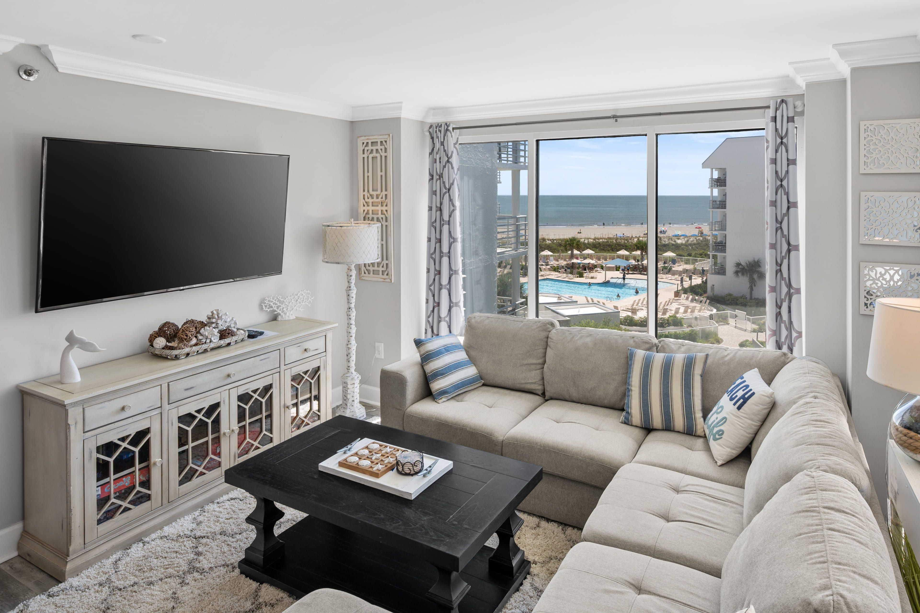 A living room with a large TV, a sofa, a coffee table, and a view of a pool and beach.