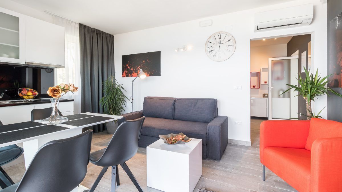 A modern living room with a grey sofa, a red sofa, a dining table with chairs, and a small white table with a vase and flowers.