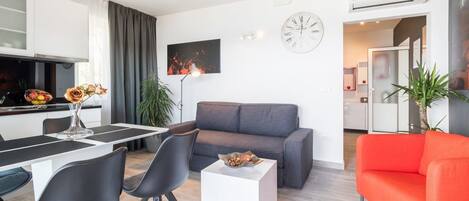 A modern living room with a grey sofa, a red sofa, a dining table with chairs, and a small white table with a vase and flowers.