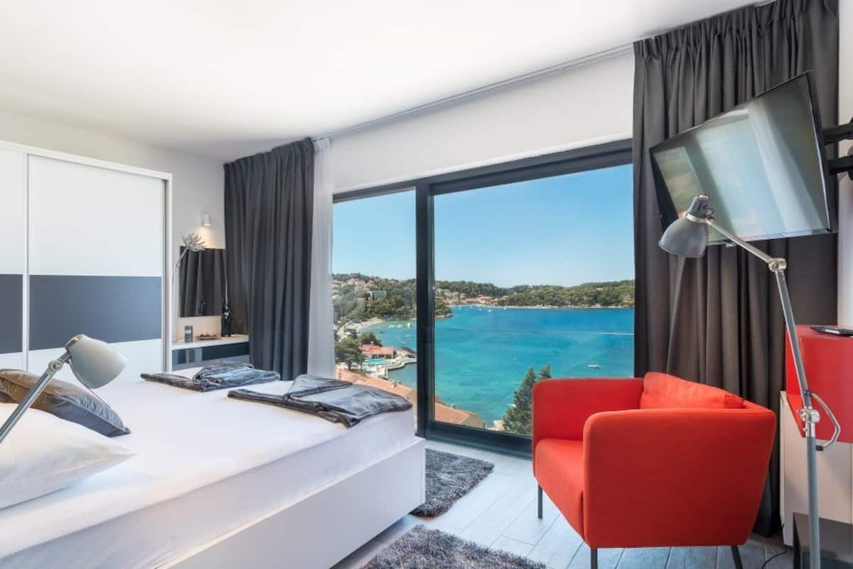 Una habitación de hotel moderna con una cama grande, una silla roja, un escritorio con una computadora y vista al mar.