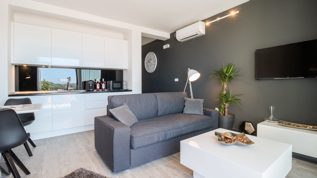 A modern living room with a grey sofa, a white coffee table, and a kitchen area with white cabinets and a large window.
