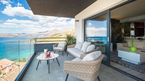 Un balcon agrémenté de meubles en osier, d’une table en verre et offrant une vue sur la mer et les montagnes.