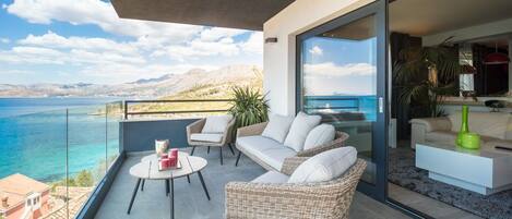 A balcony with wicker furniture, a glass table, and a view of the sea and mountains.