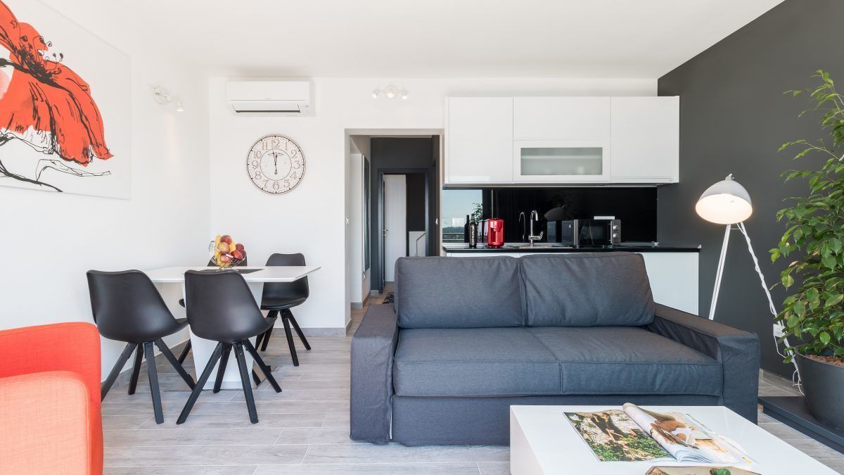 Un salon moderne avec un canapé gris, des chaises noires, une table à manger et un coin cuisine doté de meubles blancs et d’une bouilloire rouge.