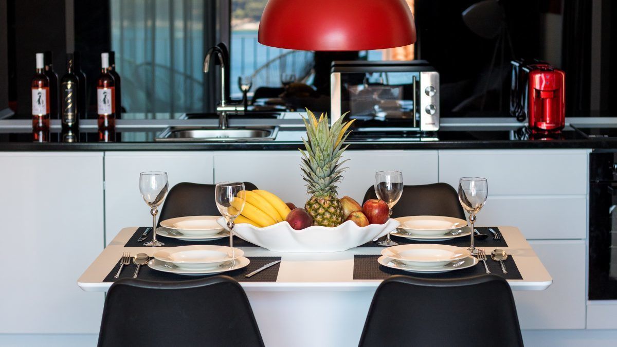 A modern kitchen with a dining table set for two, featuring a fruit bowl centerpiece, white plates, and black chairs.