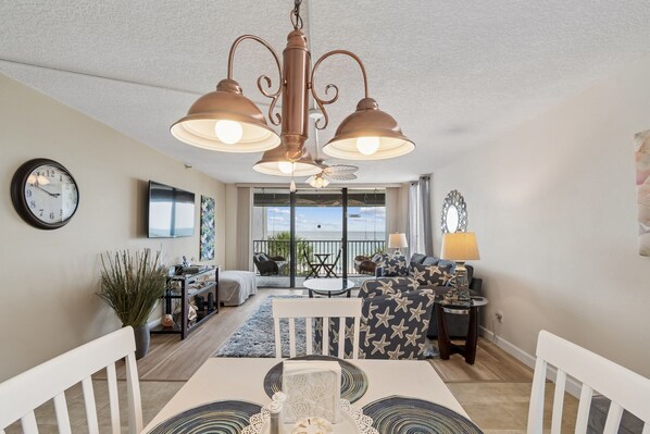 Un salon comprenant une table à manger, des chaises, un canapé, un téléviseur, une horloge et un balcon aménagé avec du mobilier d’extérieur.