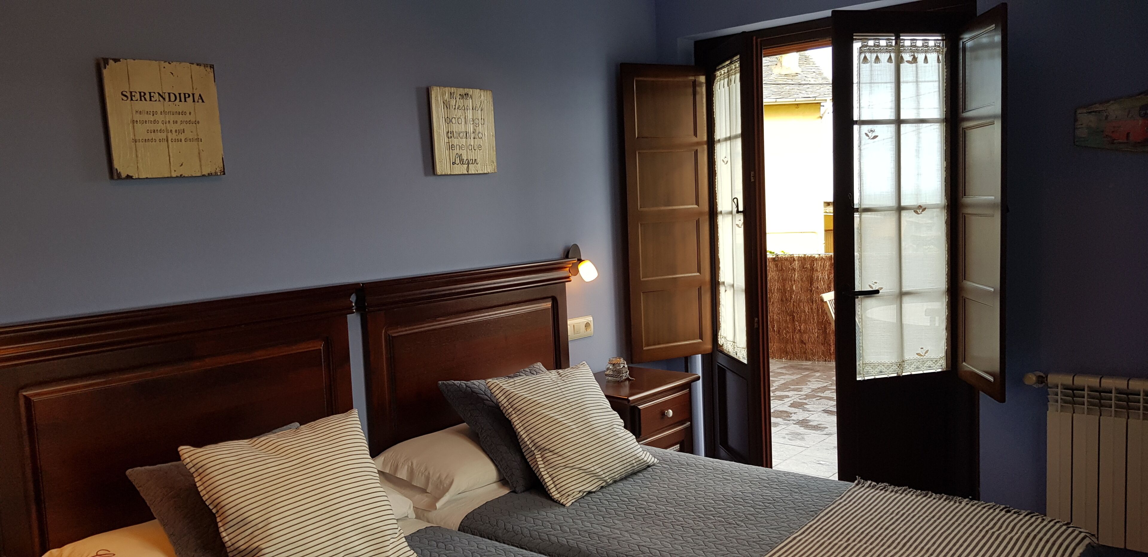 Un dormitorio con cabecera de madera, cama con almohadas a rayas y lisas, mesita de noche y ventana con vistas al exterior.
