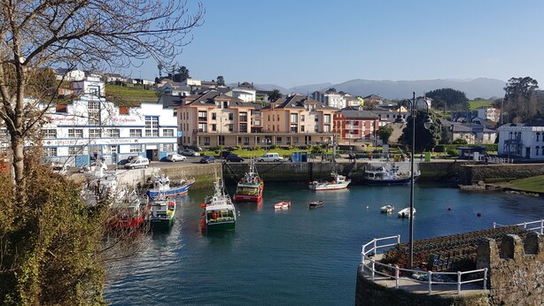 Un puerto con barcos, un edificio de varios pisos y un telón de fondo montañoso.