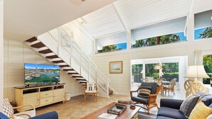 A spacious living room with a large TV, comfortable seating, a staircase, and a view of greenery outside. - SeaPines, Walk to the Beach Club, Golf view, 2 free bikes (Hilton Head Island)