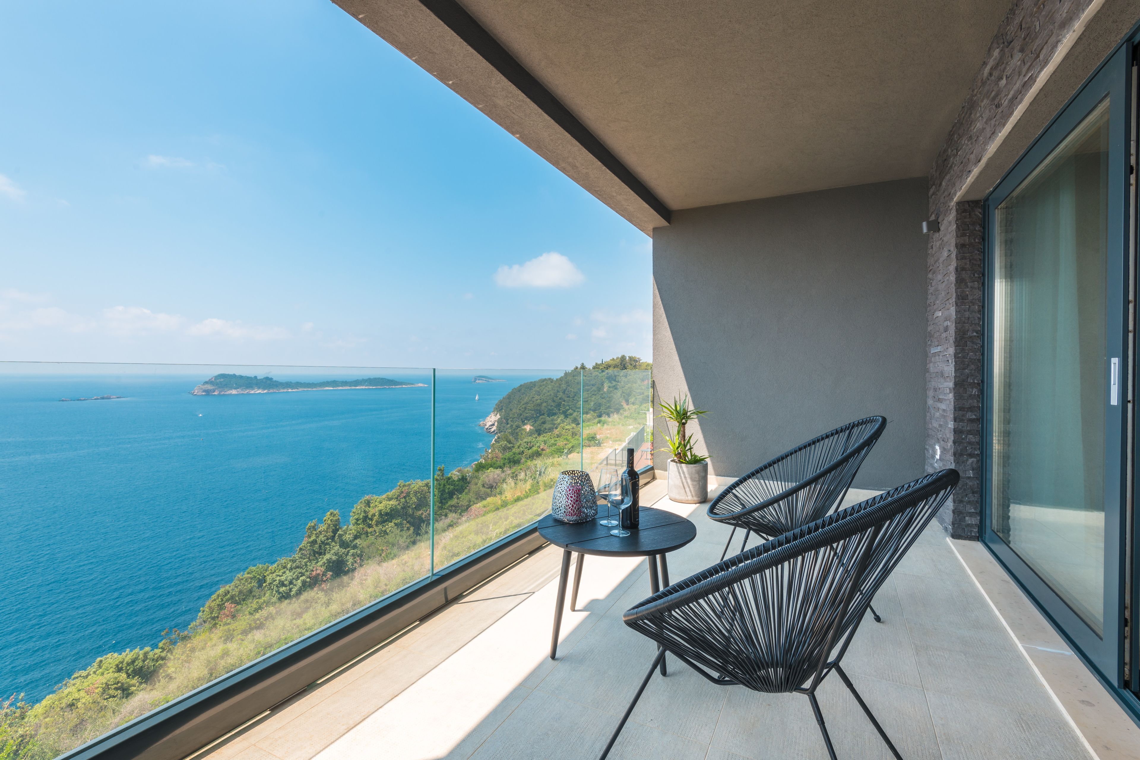A balcony with a view of the sea, two chairs, a small table with a bottle and glasses, and a potted plant.