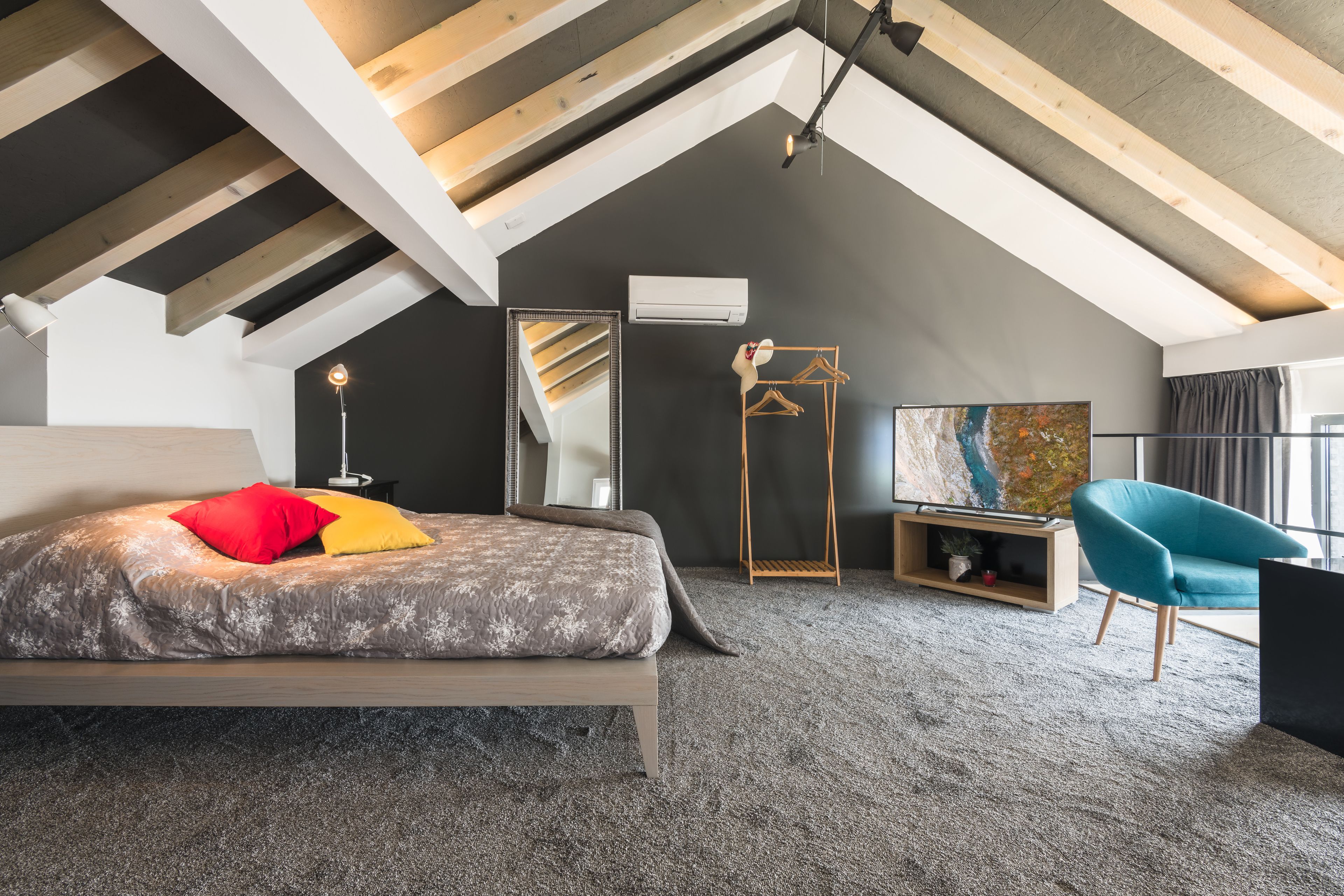 Un dormitorio con techo inclinado, una cama con ropa de cama estampada, un sillón azul y un cuadro abstracto de gran tamaño.