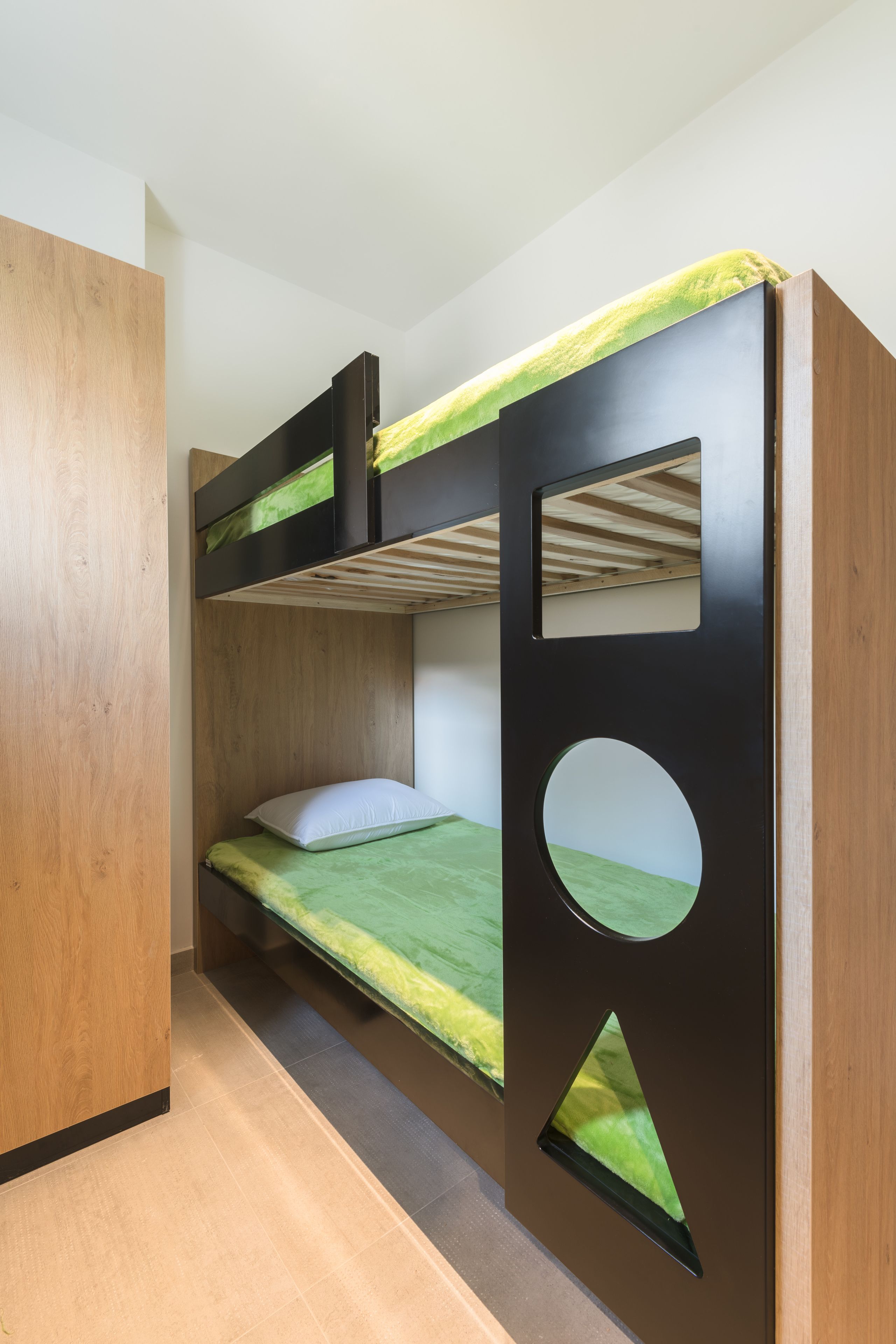 Una cama litera con colchón verde, una ventana redonda en la pared y un cabecero de madera.