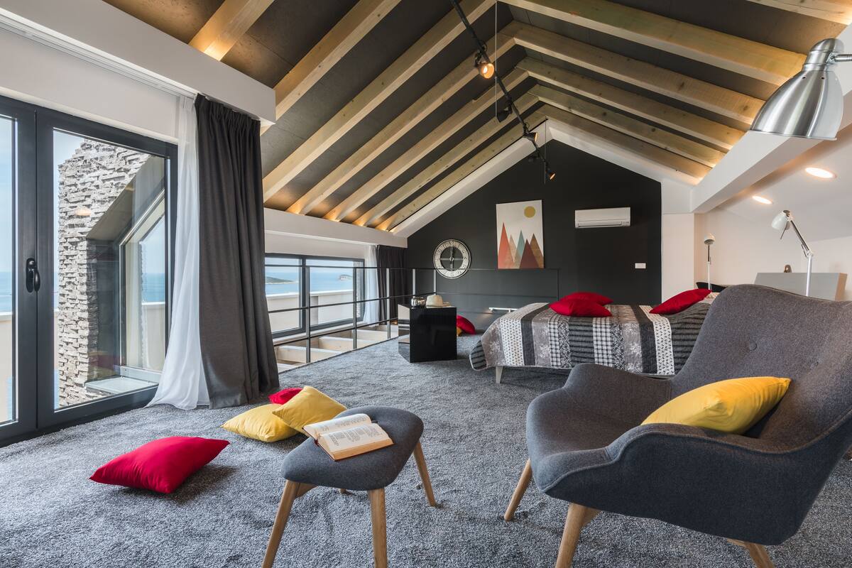 Un dormitorio moderno con techo inclinado, un ventanal grande, una cama, un sillón de lectura y una mesita.