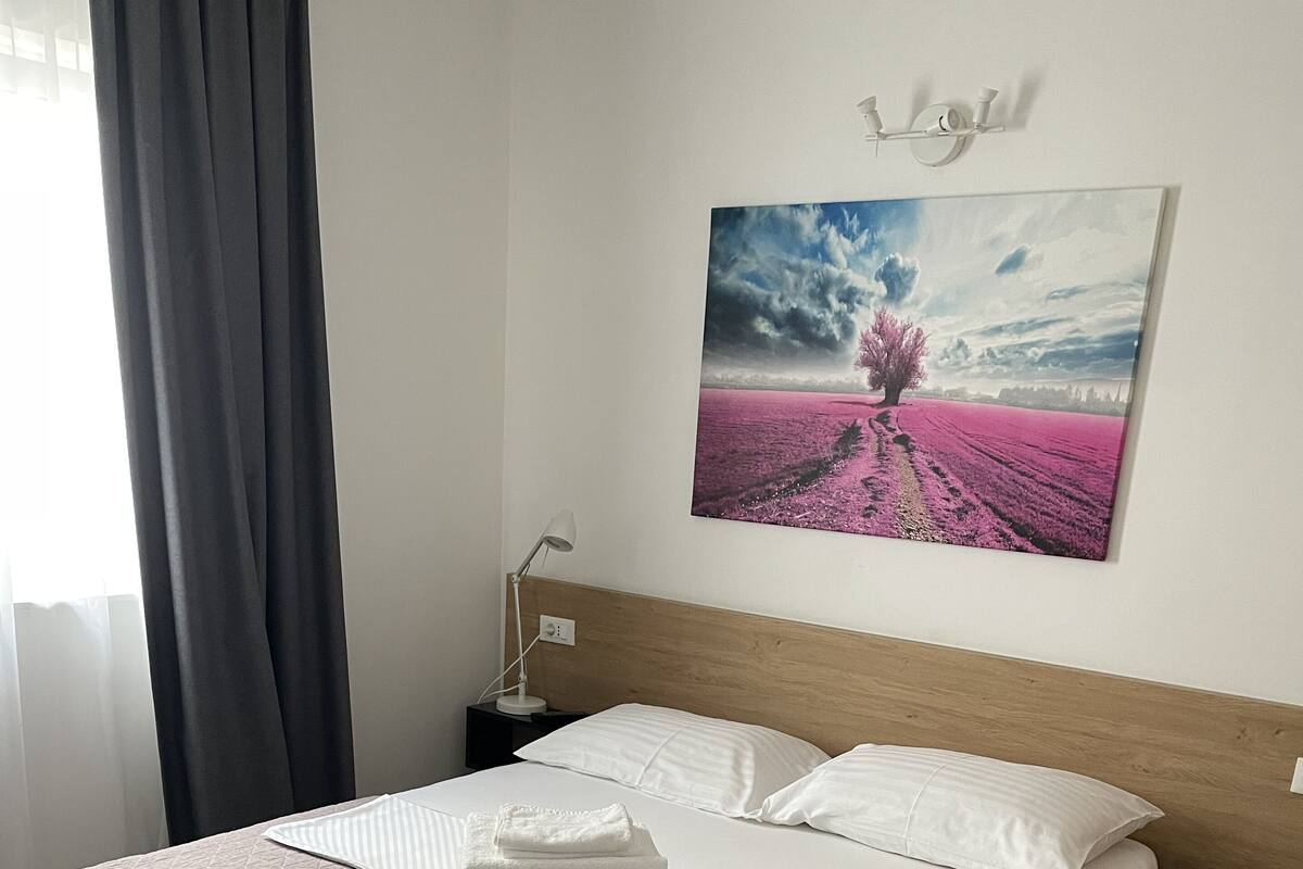 Una cama bien tendida con sábanas blancas y una toalla doblada, una lámpara de noche y un cuadro de un campo rosa en la pared.