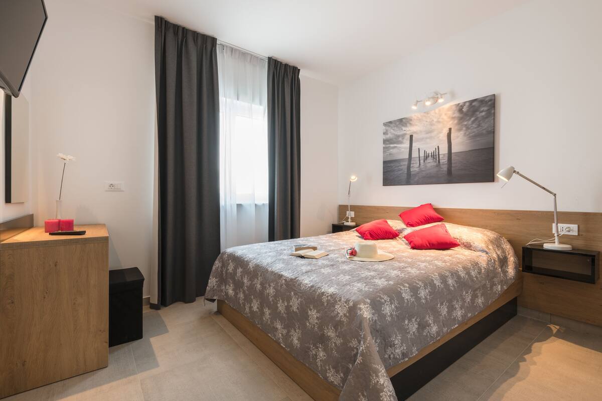 Un dormitorio moderno con una cama grande, una mesita de noche, un escritorio, una silla, una lámpara, un cuadro y una ventana con cortinas.